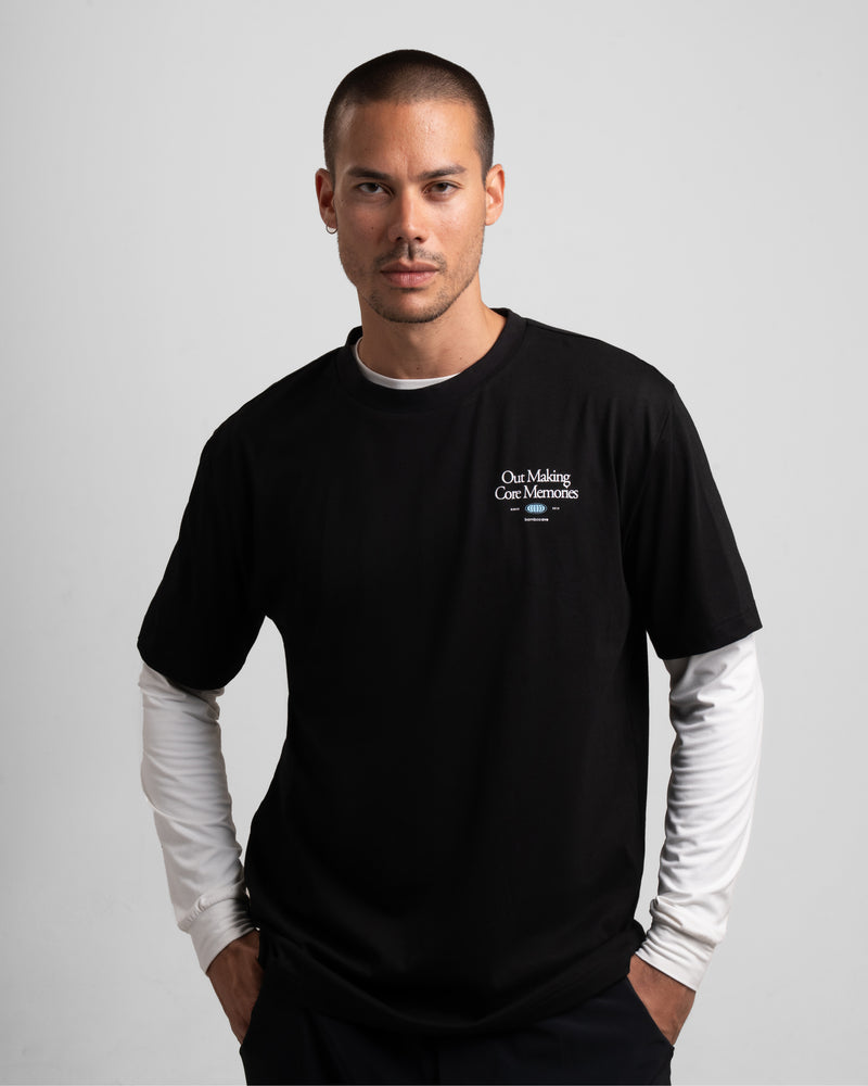 Black graphic tee featuring a small white text design, worn over a long-sleeve shirt. - Bamboo Ave